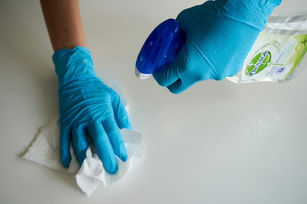 Picture of a person wearing blue gloves cleaning a white surface with spray and a paper towel
