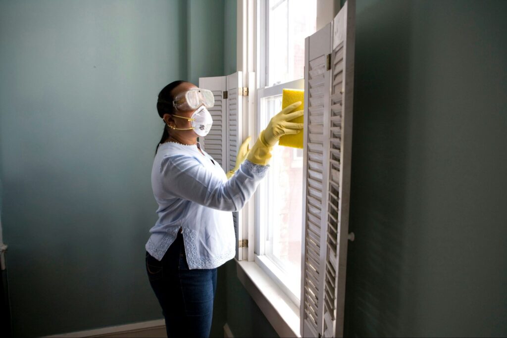 Picture of a woman wearing protective goggles, a mask, and yellow gloves cleaning window shutters