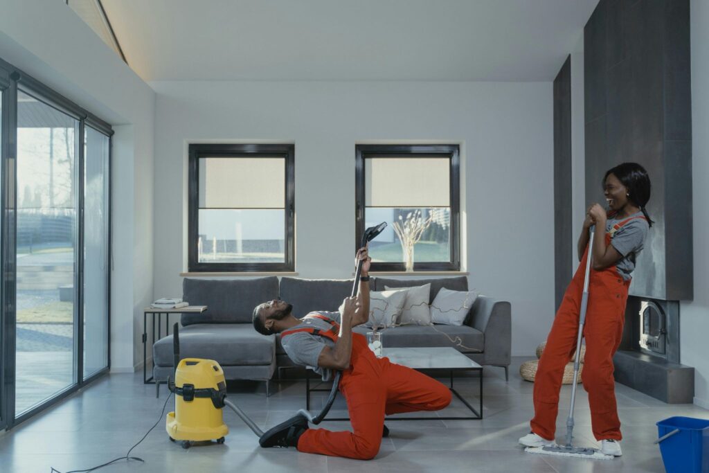 Picture of a pair of cleaners in orange overalls having fun and playing air guitar with a vacuum
