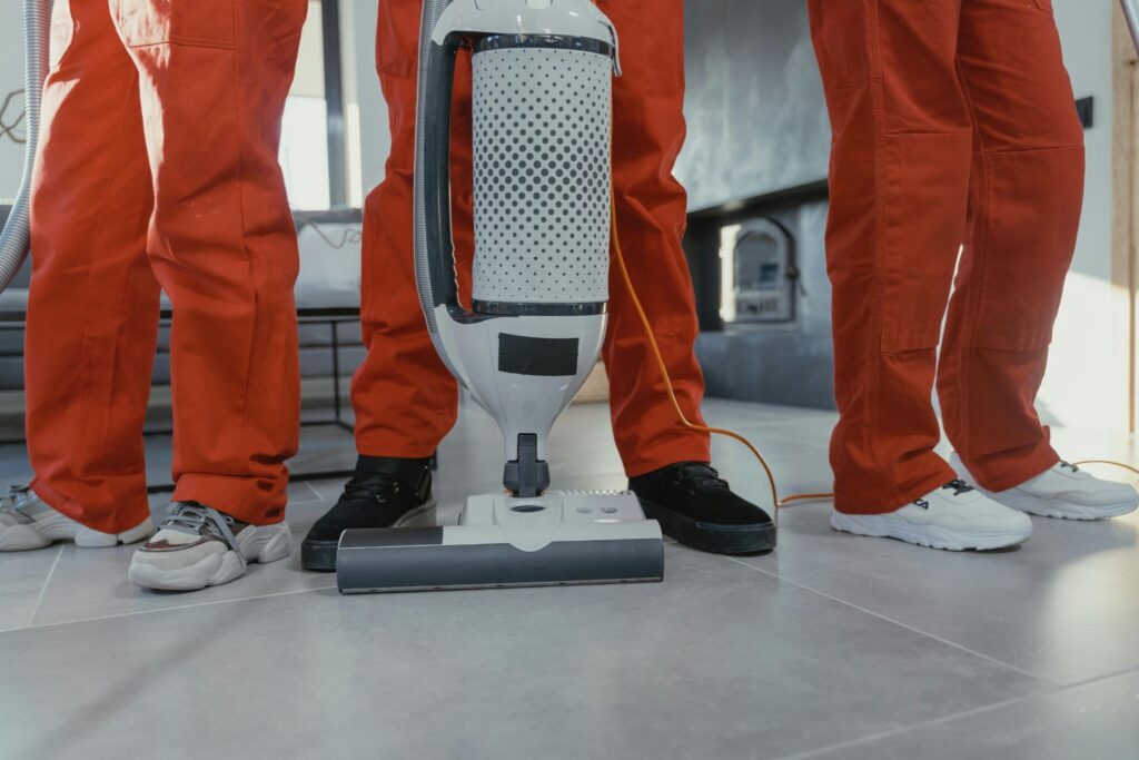 Picture of a vacuum cleaner on a tiled floor surrounded by cleaners in orange overalls