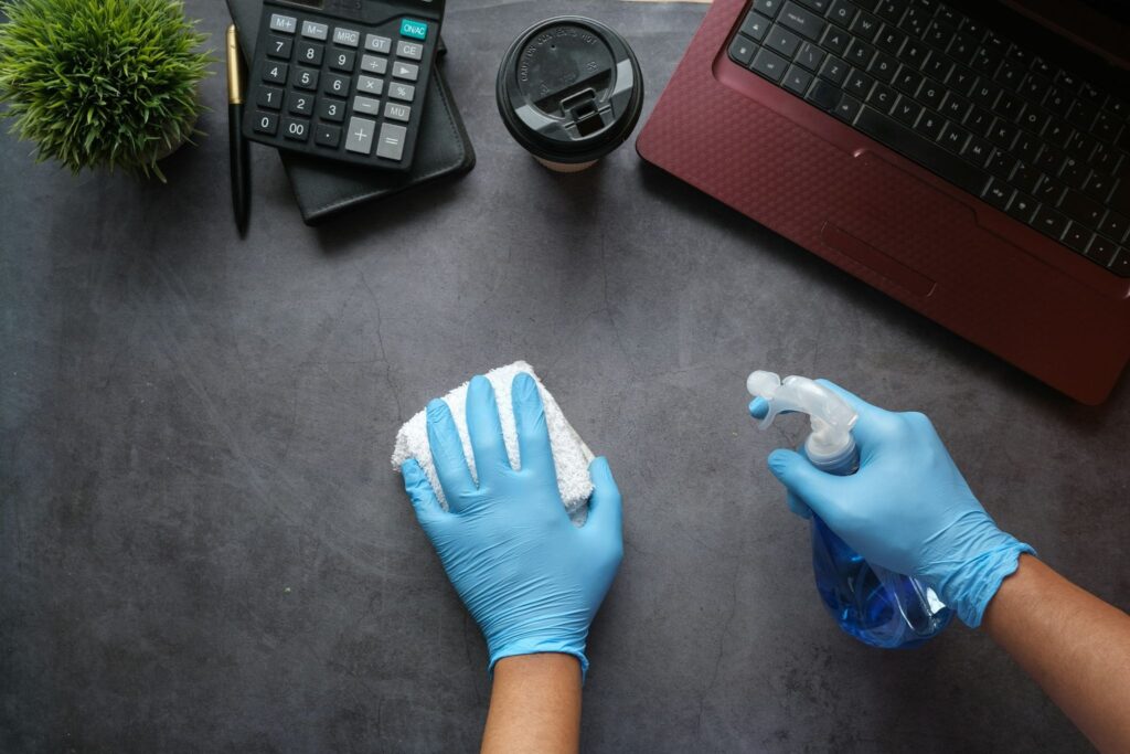 Picture of a top down view of blue gloved hands cleaning a desk next to a laptop