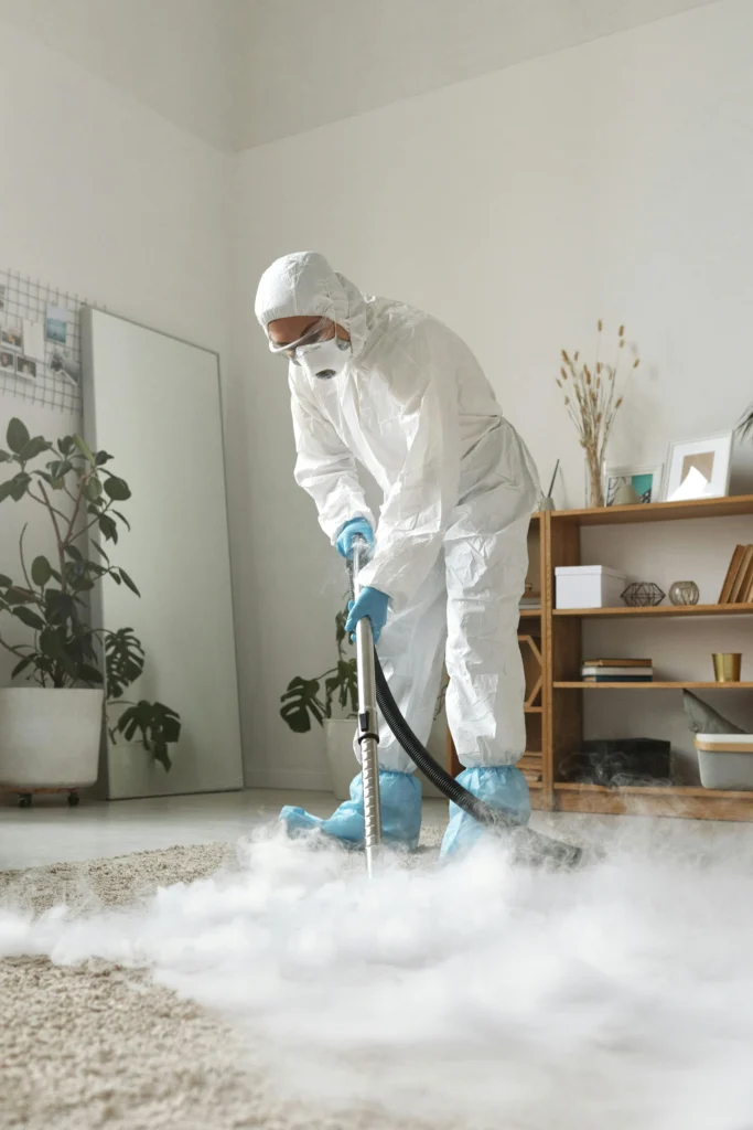 picture of a cleaner in a full white protective suit and mask steam cleaning a living room carpet