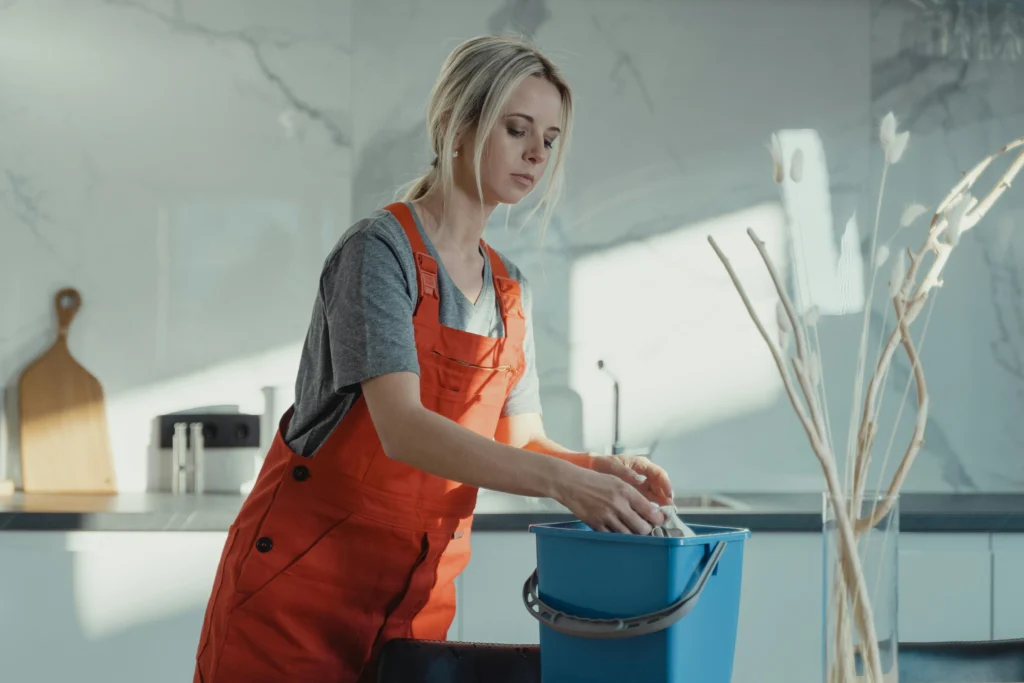 picture of a female cleaner in orange overalls placing a cloth into a blue bucket in a sunny kitchen