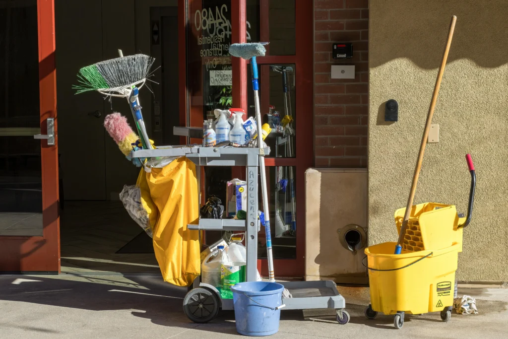 picture of a fully loaded grey cleaning cart alongside a yellow mop bucket parked near a building entrance