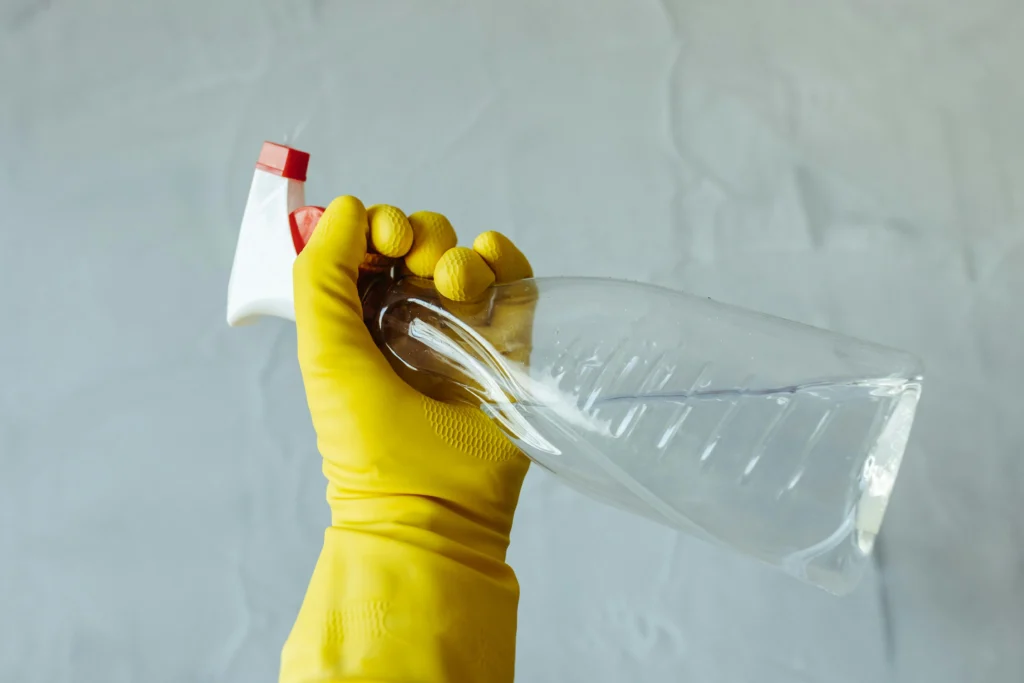 picture of a hand in a yellow rubber glove holding a clear spray bottle against a textured grey background