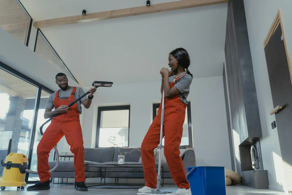 picture of a man and woman in orange overalls playfully using cleaning tools as musical instruments in a living room
