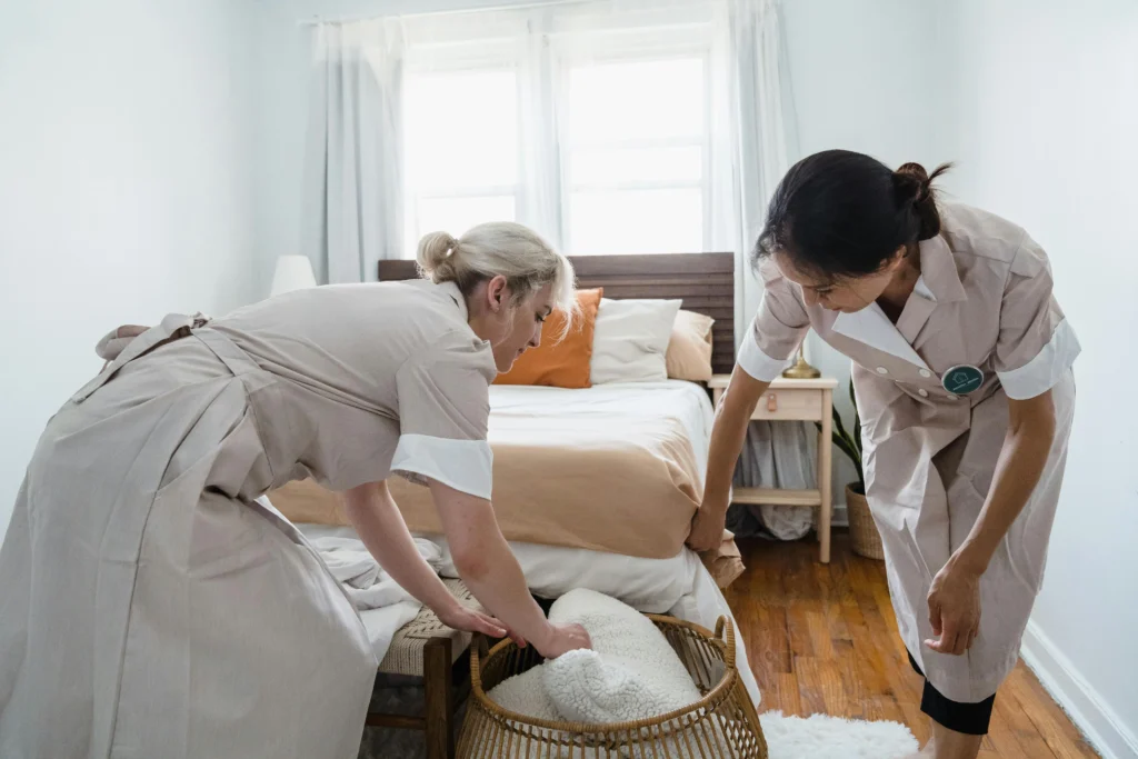 picture of a pair of female cleaners in beige uniforms arranging bedding and a woven basket at the foot of a bed