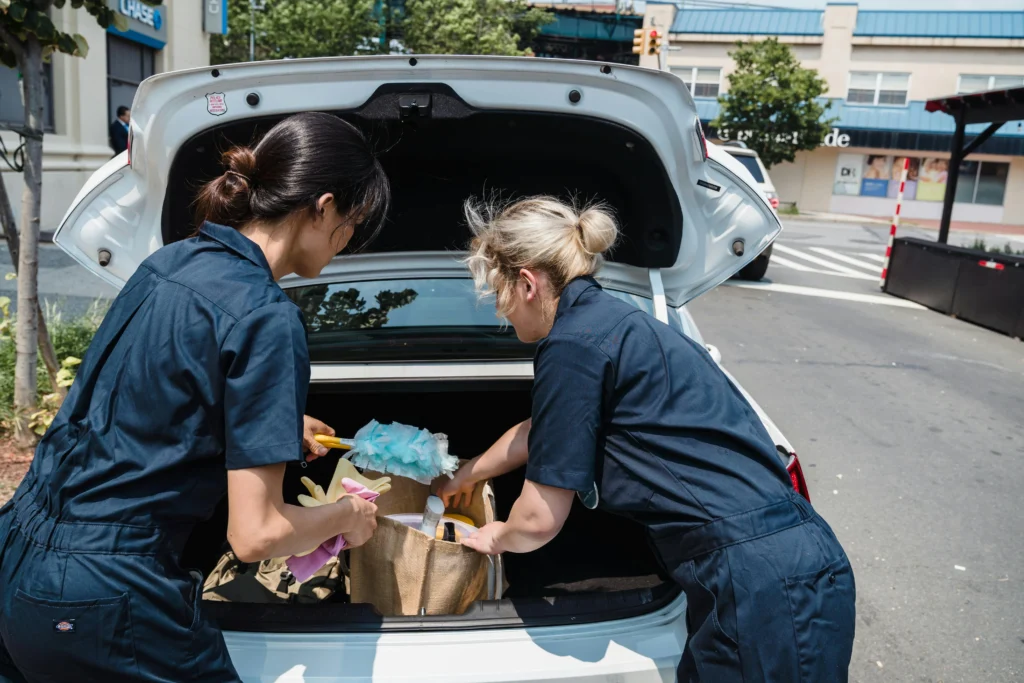 picture of a pair of female cleaners in dark blue uniforms retrieving supplies from the open trunk of a white car