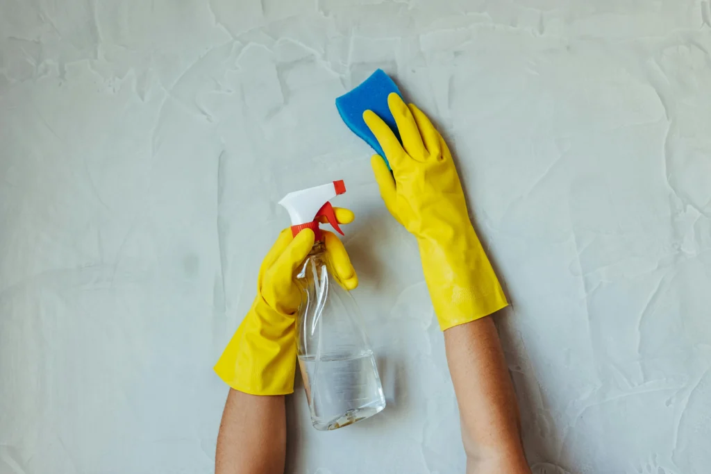 picture of a person wearing yellow rubber gloves holding a spray bottle and blue sponge against a grey wall