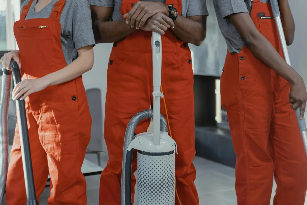 picture of the midsection of three cleaners in orange overalls standing with a white vacuum