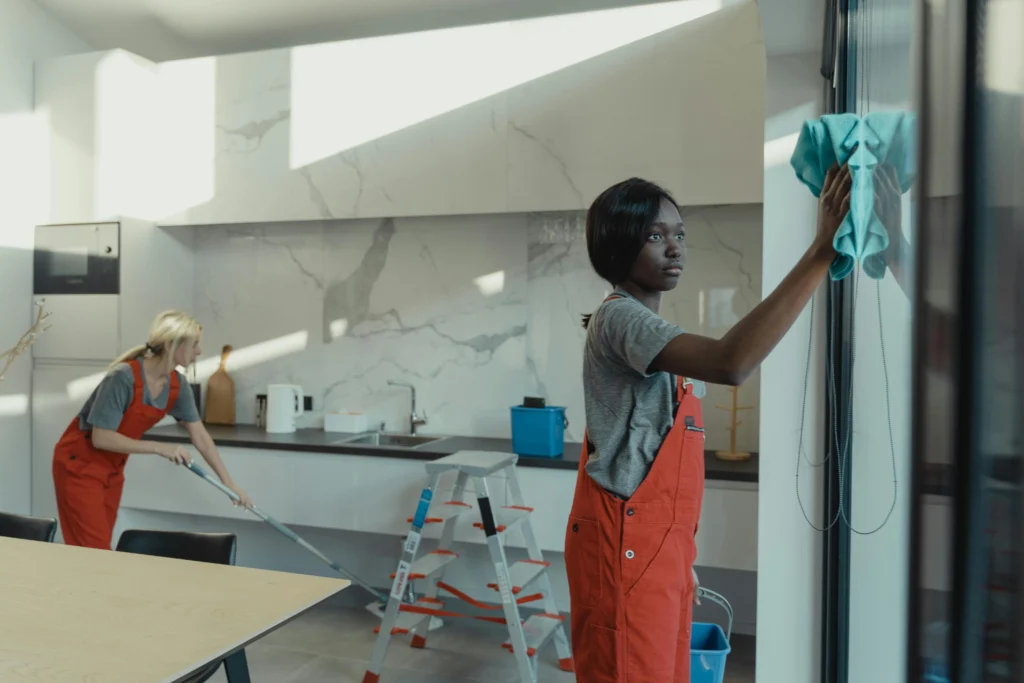 picture of two cleaners in orange uniforms working in a kitchen while one wipes a glass door