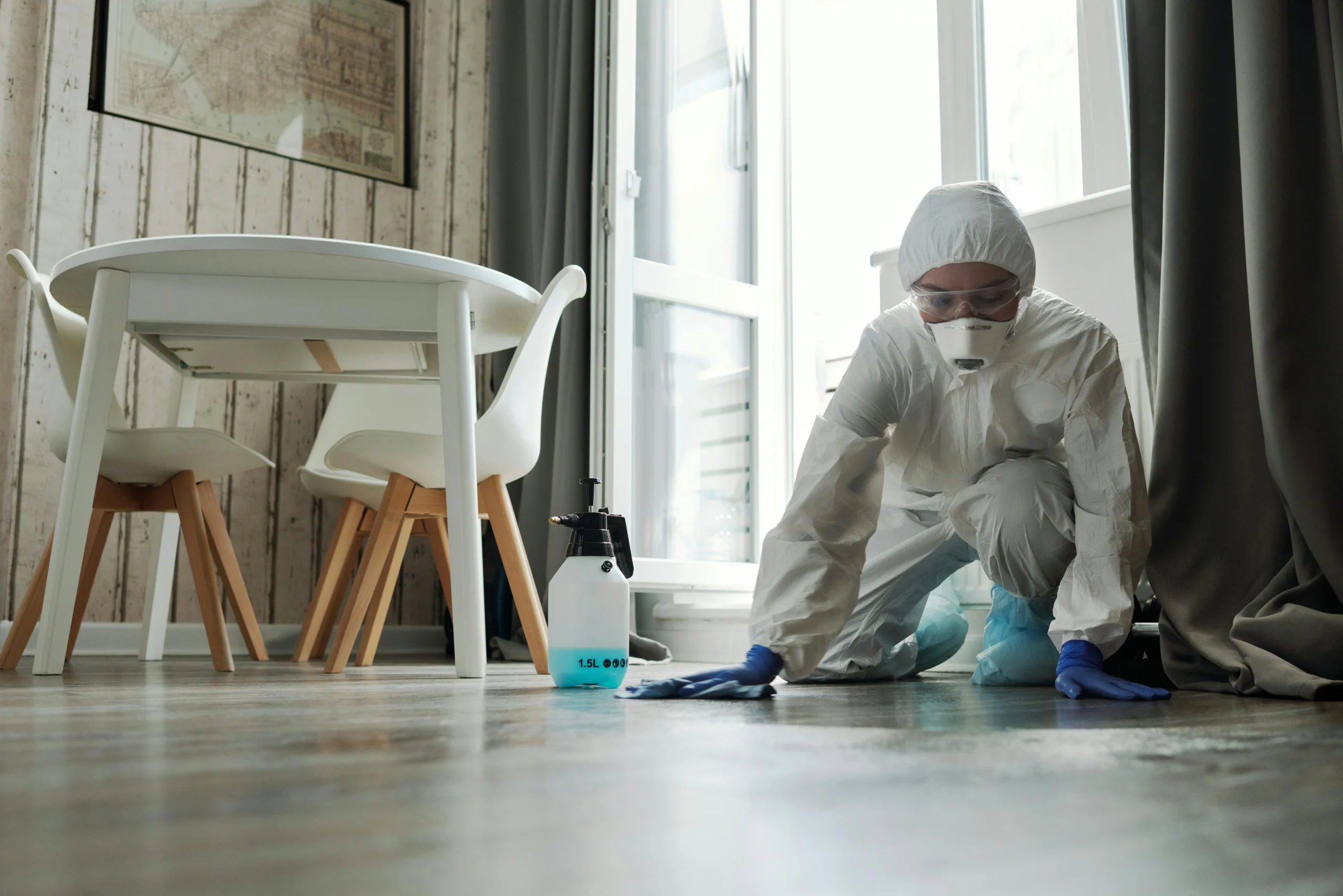 Picture of a cleaner wearing a white hazmat suit while holding a spray bottle and colorful cloths.