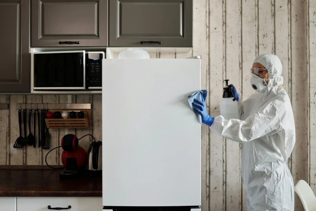 Picture of a cleaner in a white protective suit spraying and wiping a white refrigerator