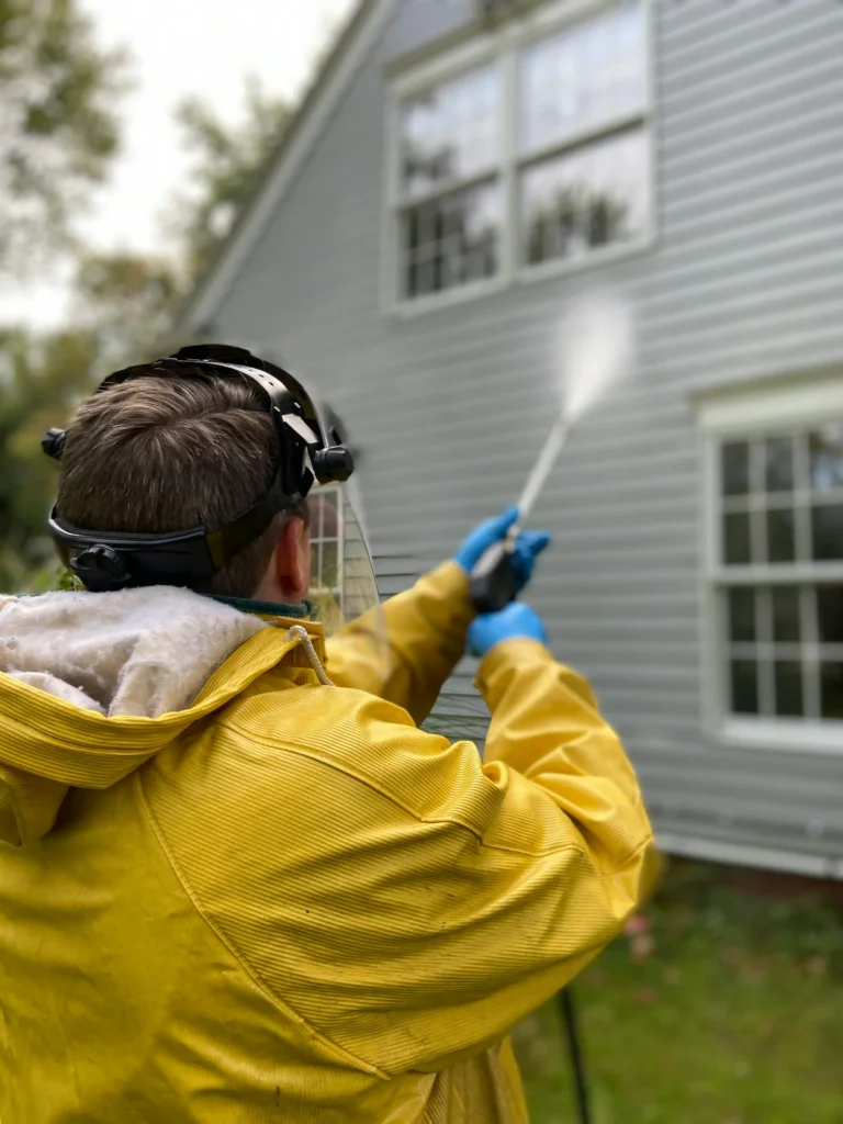 Picture of a person wearing a yellow rain jacket and safety gear spraying the exterior siding of a house