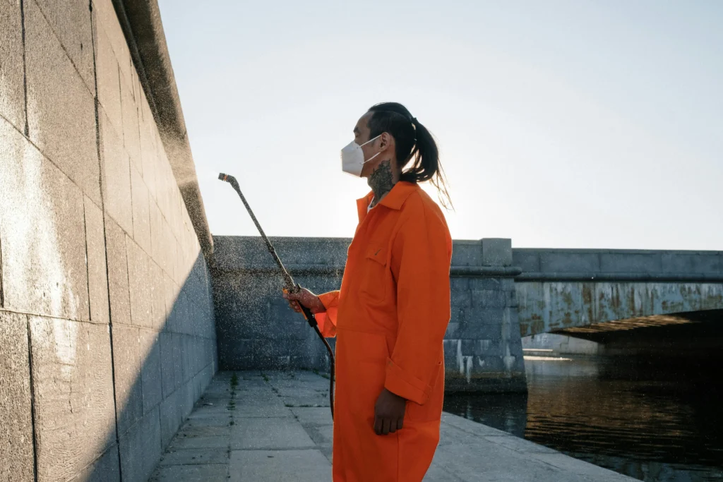 Picture of a worker in an orange protective suit and mask spraying a concrete wall outdoors near a body of water