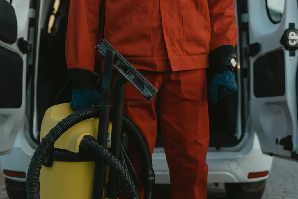 Picture of a cleaner in red overalls holding a vacuum by a van